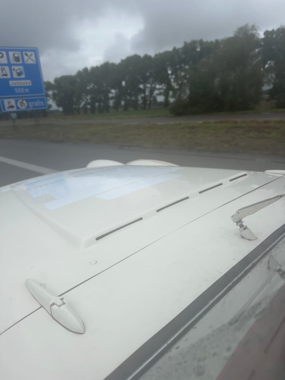 Looking along TS2's wet bonnet on the E40, a blue Belgian services sign at the roadside reading Jabbeke 500 m