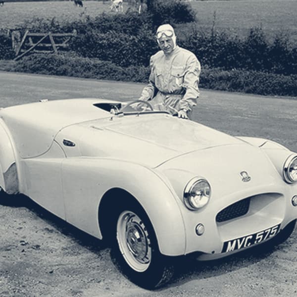 Ken Richardson in white overalls and helmet standing beside MVC 575, the streamlined pre-production TR2, on the Jabbeke road in 1953