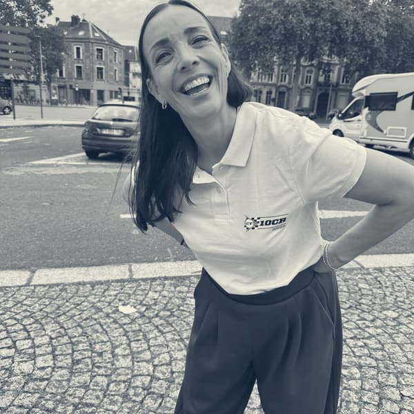 Evie laughing with her head tilted, in a cream 10CR polo shirt, on a cobbled square with parked cars and town houses behind