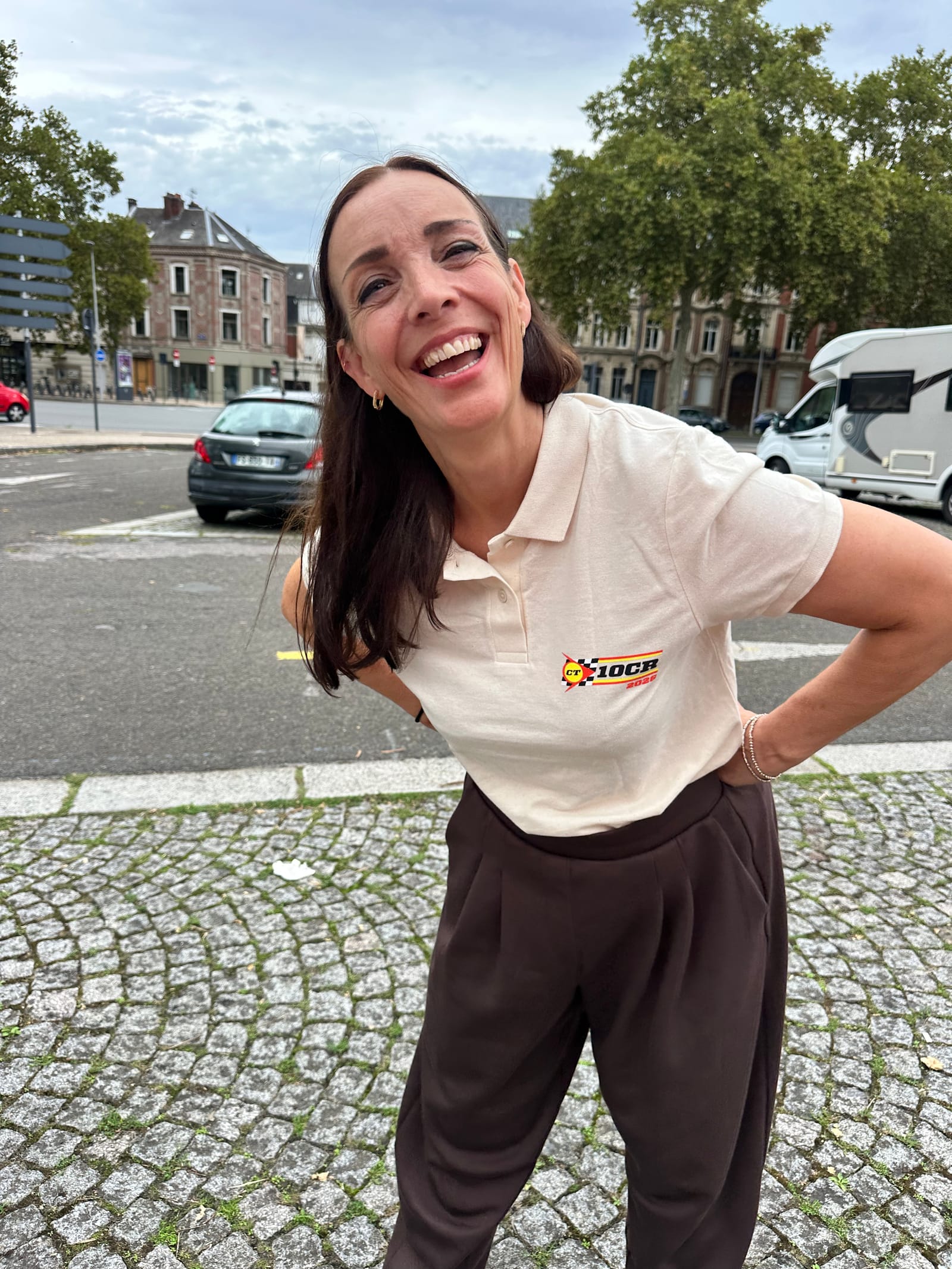 Evie laughing with her head tilted, in a cream 10CR polo shirt, on a cobbled square with parked cars and town houses behind