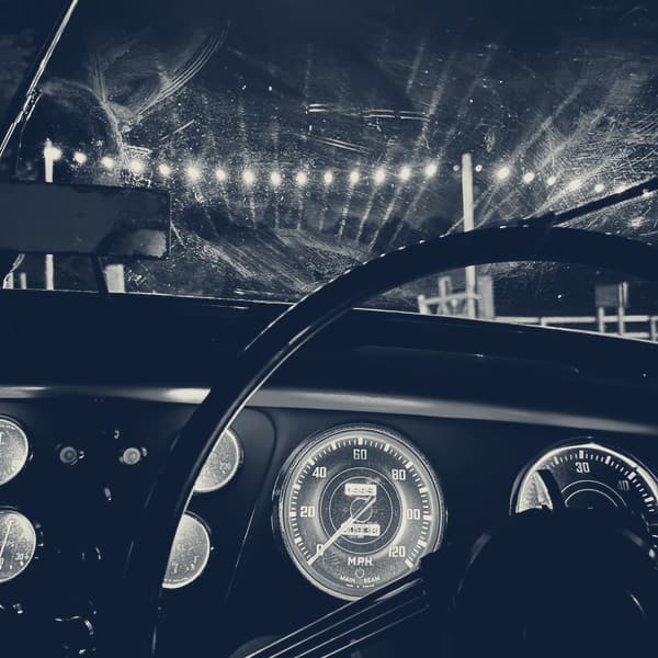 From the driver's seat at night: the wheel, the lit gauges and a windscreen smeared by the wipers, with the pub's string lights beyond