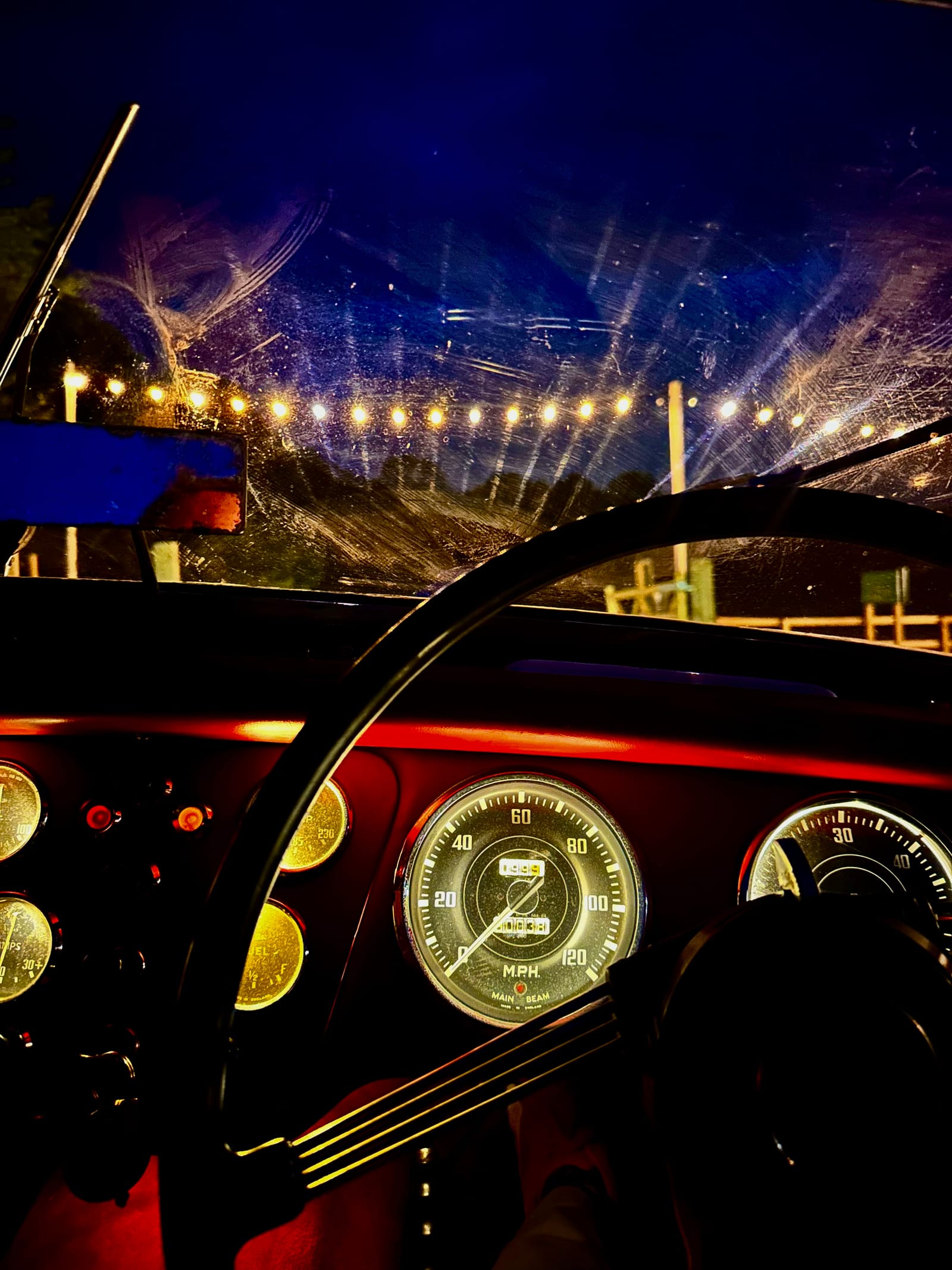 From the driver's seat at night: the wheel, the lit gauges and a windscreen smeared by the wipers, with the pub's string lights beyond
