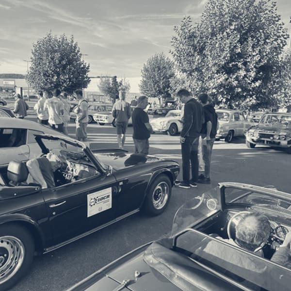 A supermarket car park full of Triumphs in early morning sun, TR6s and saloons in the bays with people standing about between them