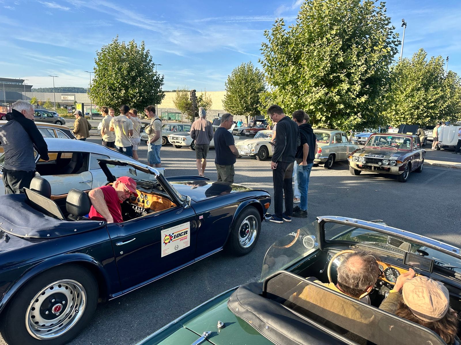 A supermarket car park full of Triumphs in early morning sun, TR6s and saloons in the bays with people standing about between them