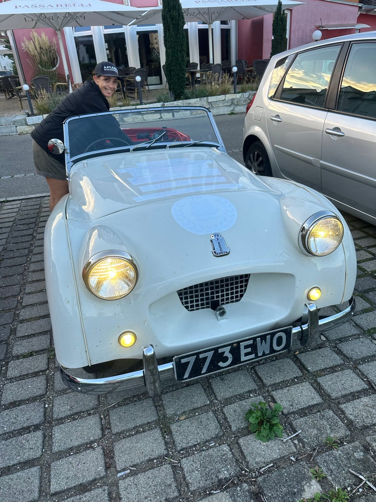Evie leaning over TS2's scuttle outside a restaurant at dusk, grinning, with both headlights and sidelights lit and 773 EWO on the plate