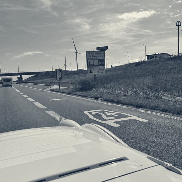 Motorway seen over TS2's white scuttle, a blue sign for the aire and Luxembourg 6 km, wind turbines on the ridge beyond