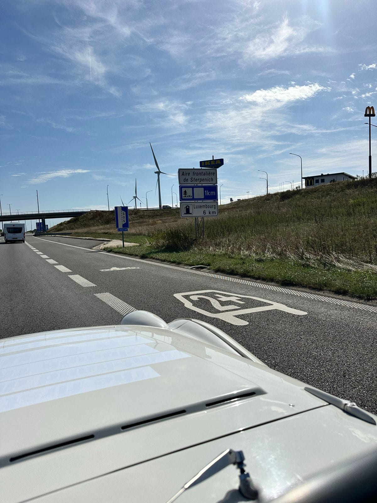 Motorway seen over TS2's white scuttle, a blue sign for the aire and Luxembourg 6 km, wind turbines on the ridge beyond