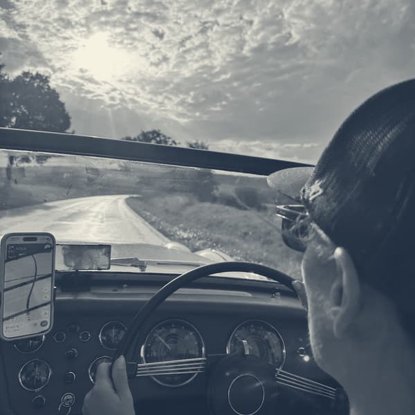 Evie at the wheel in a cap, hands on the rim, low sun through TS2's windscreen and a phone showing the route propped on the dashboard