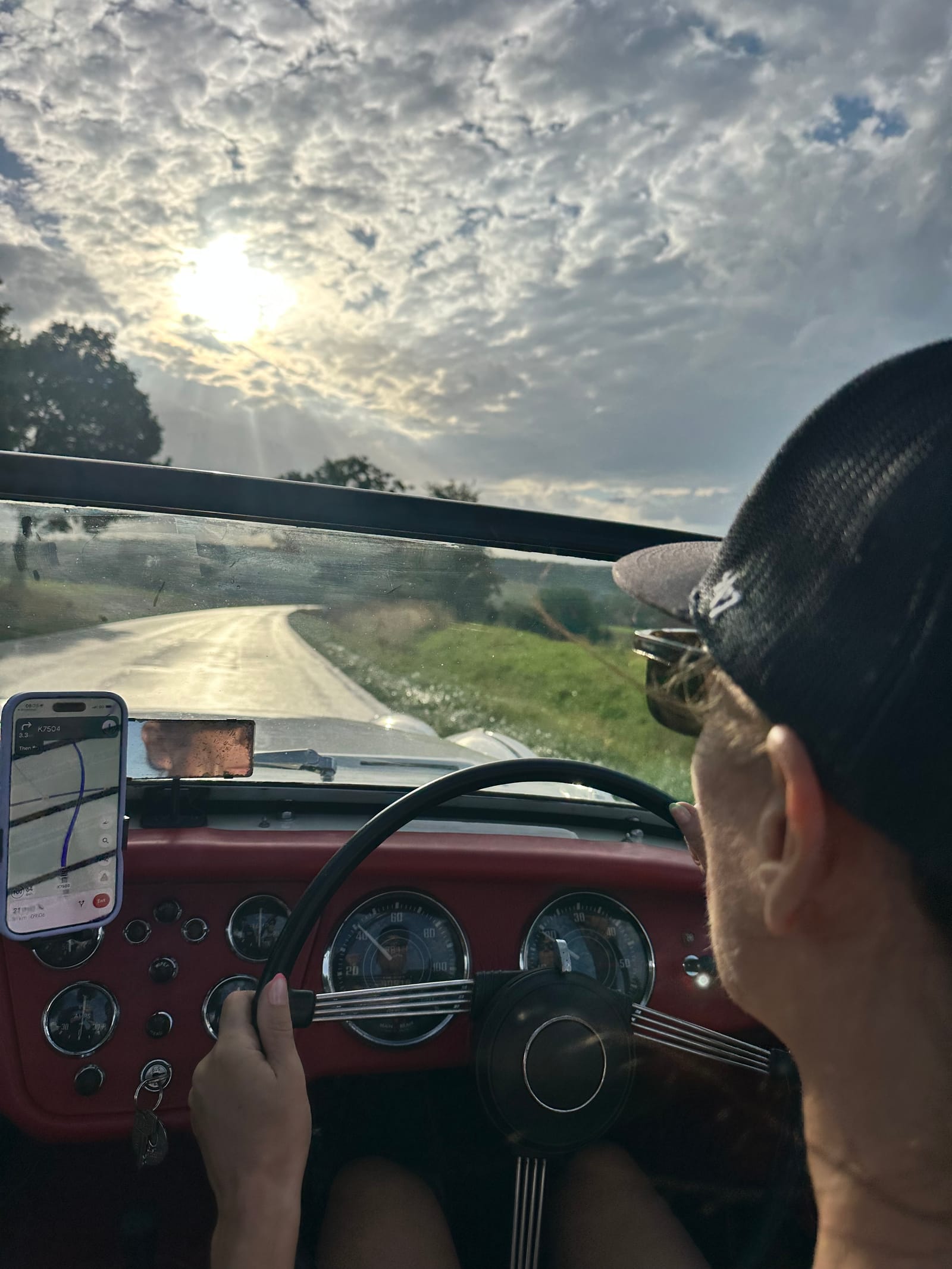 Evie at the wheel in a cap, hands on the rim, low sun through TS2's windscreen and a phone showing the route propped on the dashboard