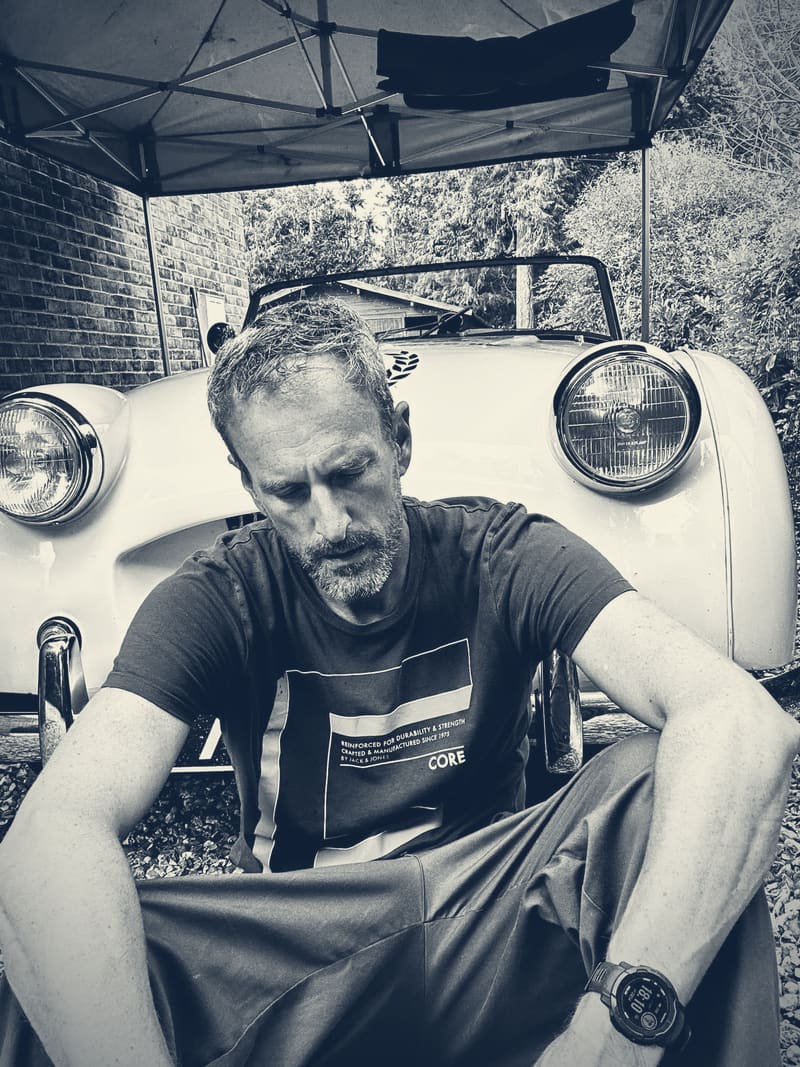 Andy sitting on the gravel in front of TS2 under a gazebo, head down, forearms on his knees, in black and white