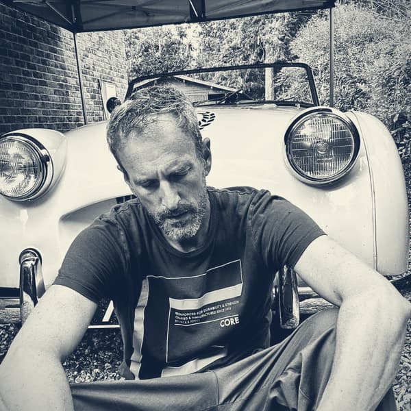 Andy sitting on the gravel in front of TS2 under a gazebo, head down, forearms on his knees, in black and white