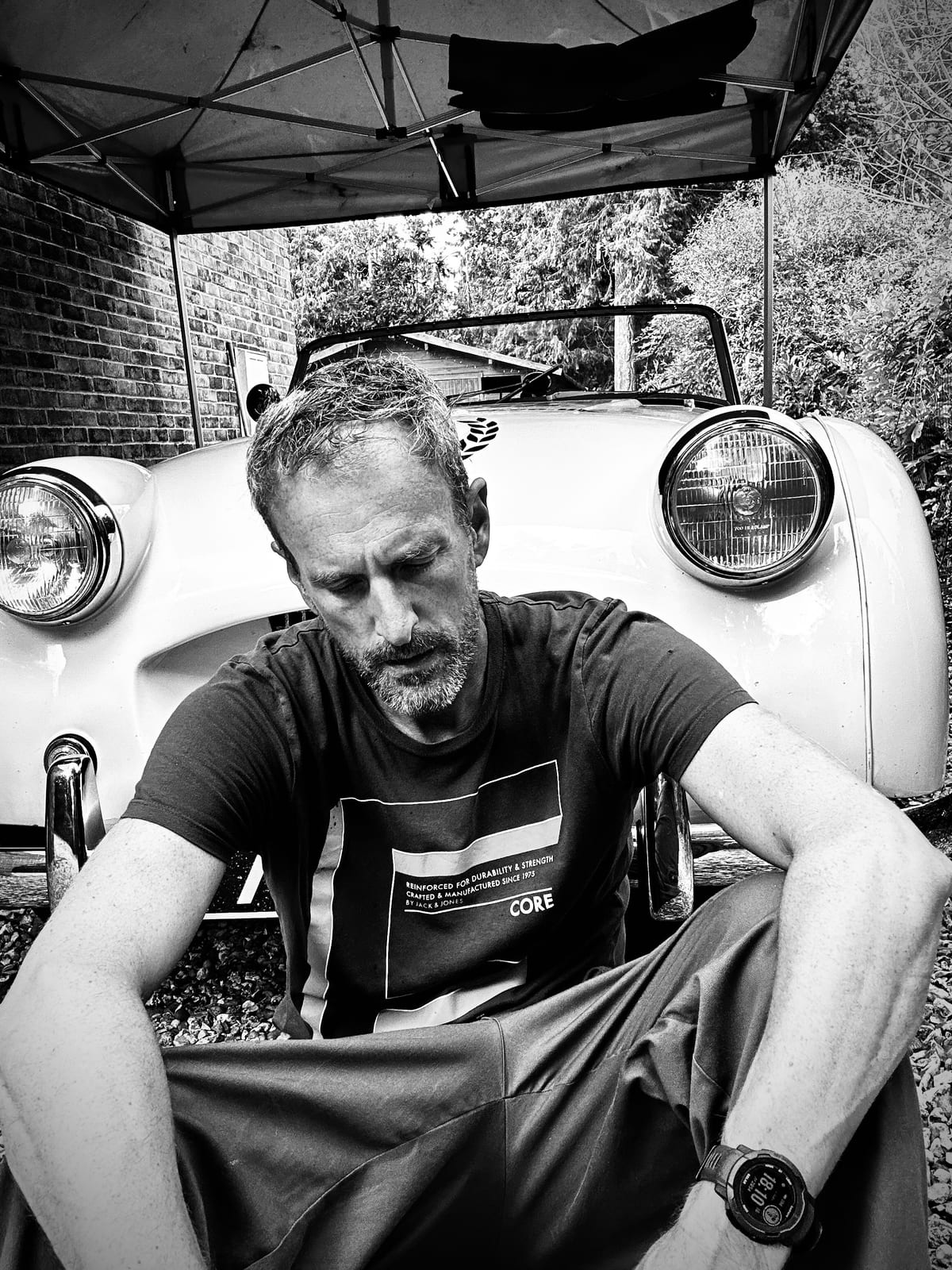 Andy sitting on the gravel in front of TS2 under a gazebo, head down, forearms on his knees, in black and white