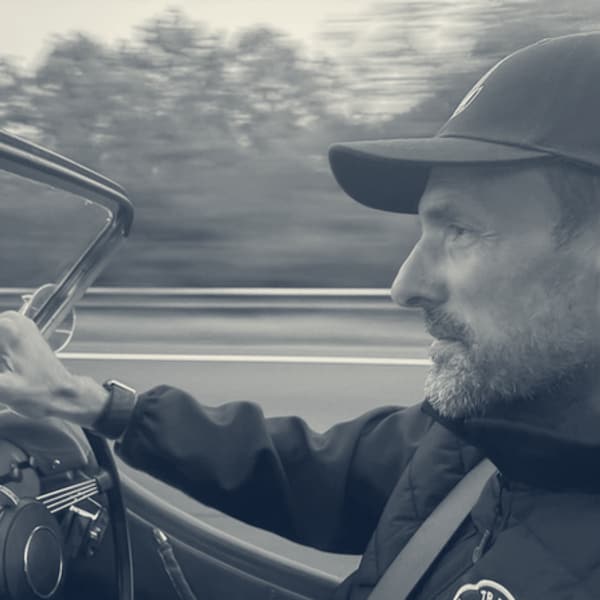 Andy at the wheel in a cap, one hand on the rim, the lit rev counter below and the trees going by in a blur
