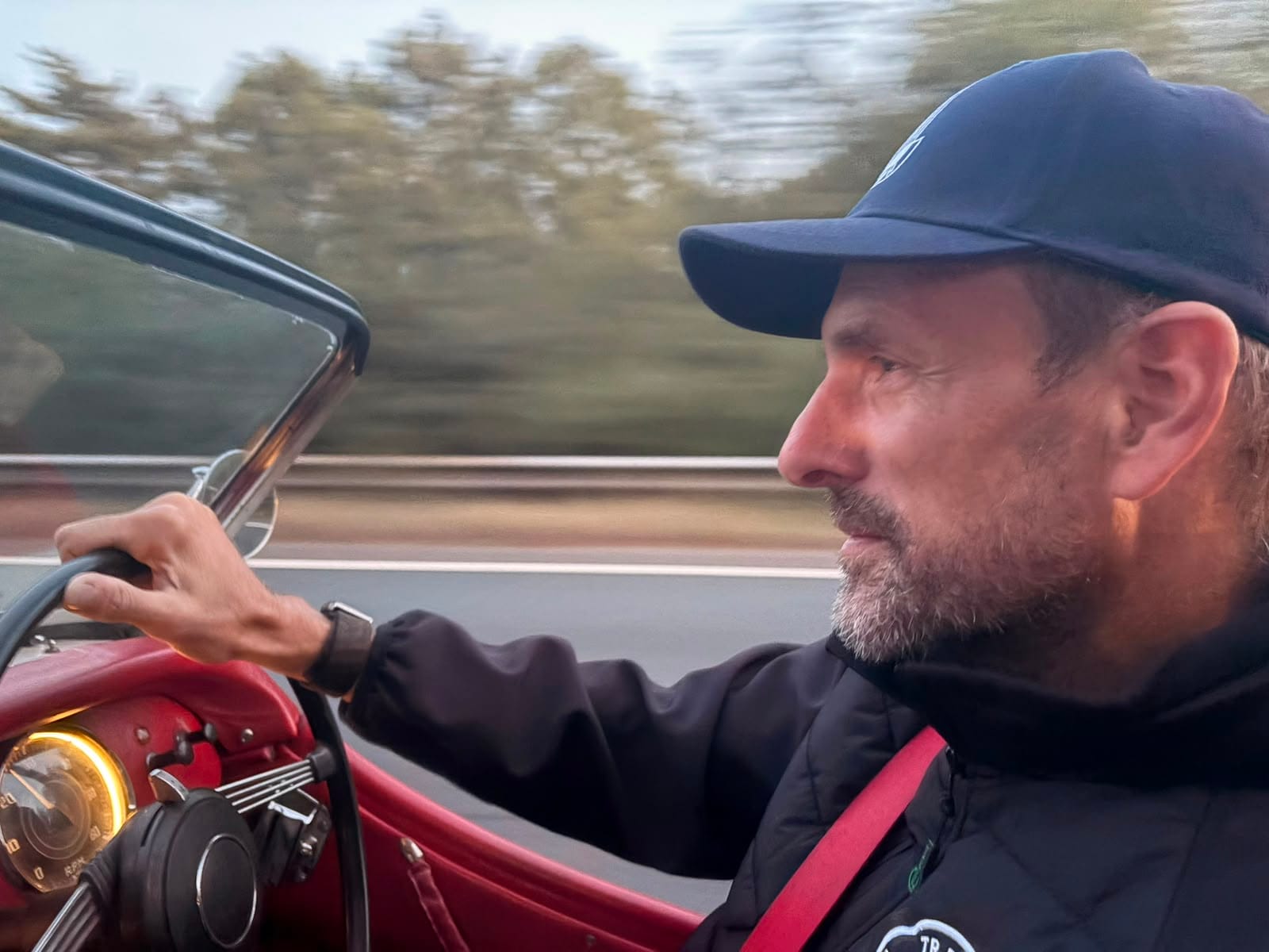 Andy at the wheel in a cap, one hand on the rim, the lit rev counter below and the trees going by in a blur