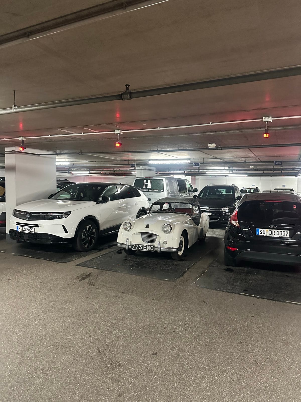 TS2 in an underground car park, wedged nose out between two modern SUVs, wet paint under the strip lights