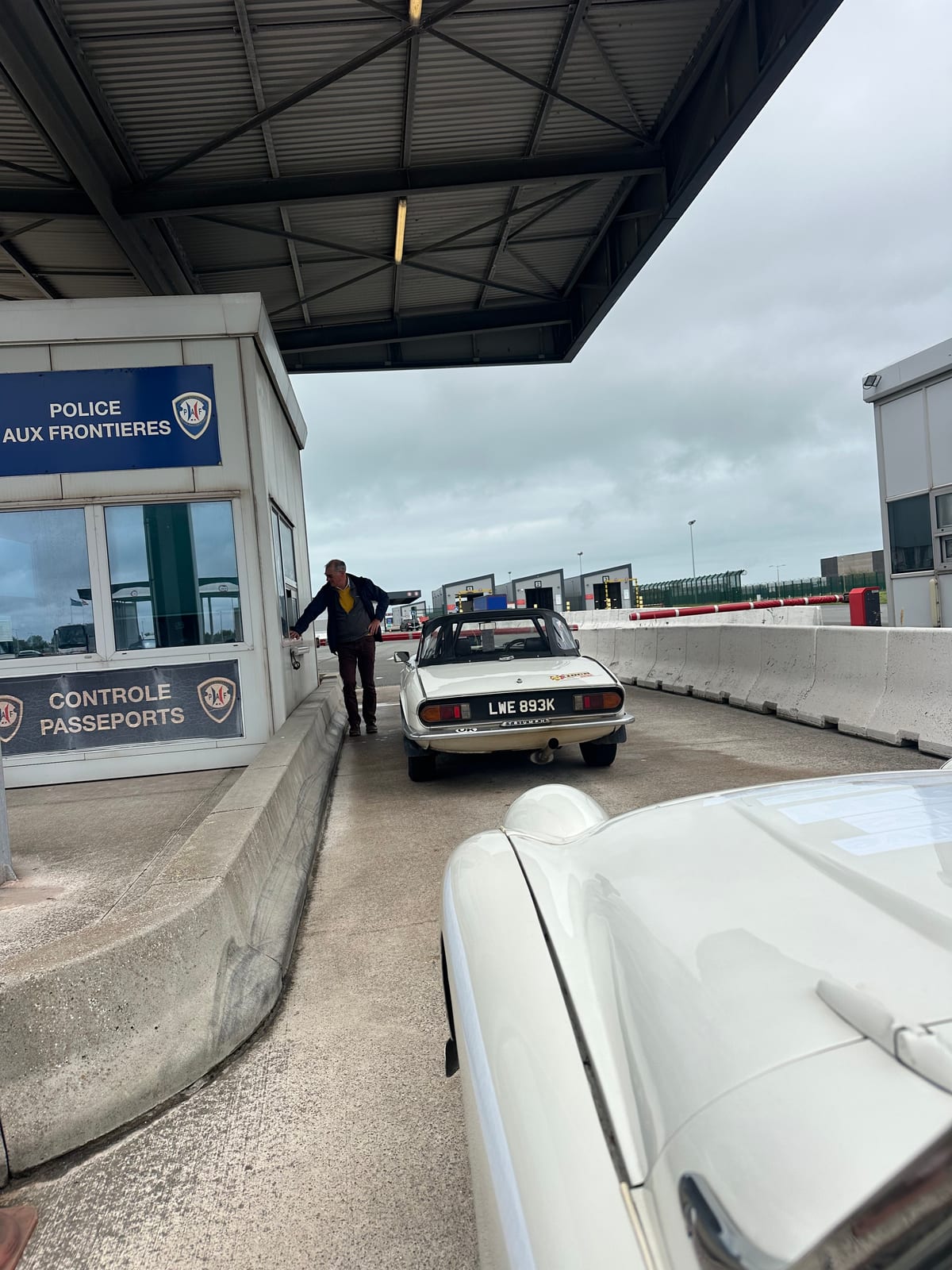 A Police aux Frontieres booth at Dunkirk marked Controle Passeports, another entrant's Triumph saloon at the window ahead, TS2's white wing in the corner
