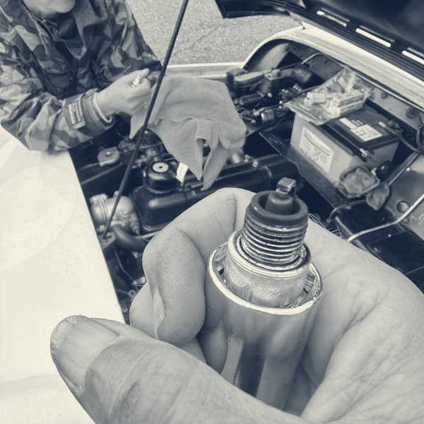 A spark plug held up to the camera, its electrode black with soot, Evie grinning over the open bonnet behind