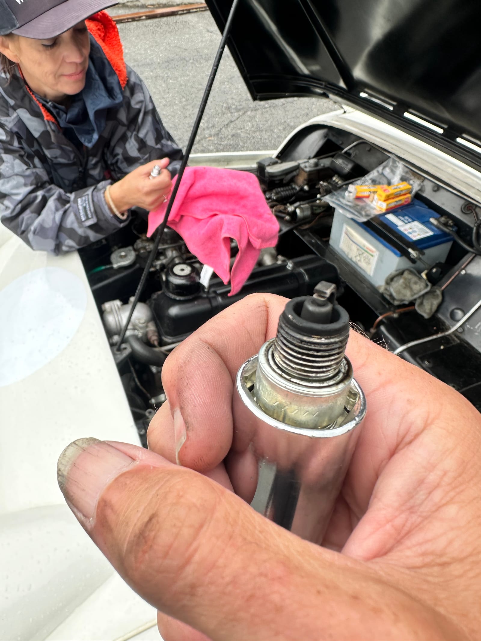 A spark plug held up to the camera, its electrode black with soot, Evie grinning over the open bonnet behind
