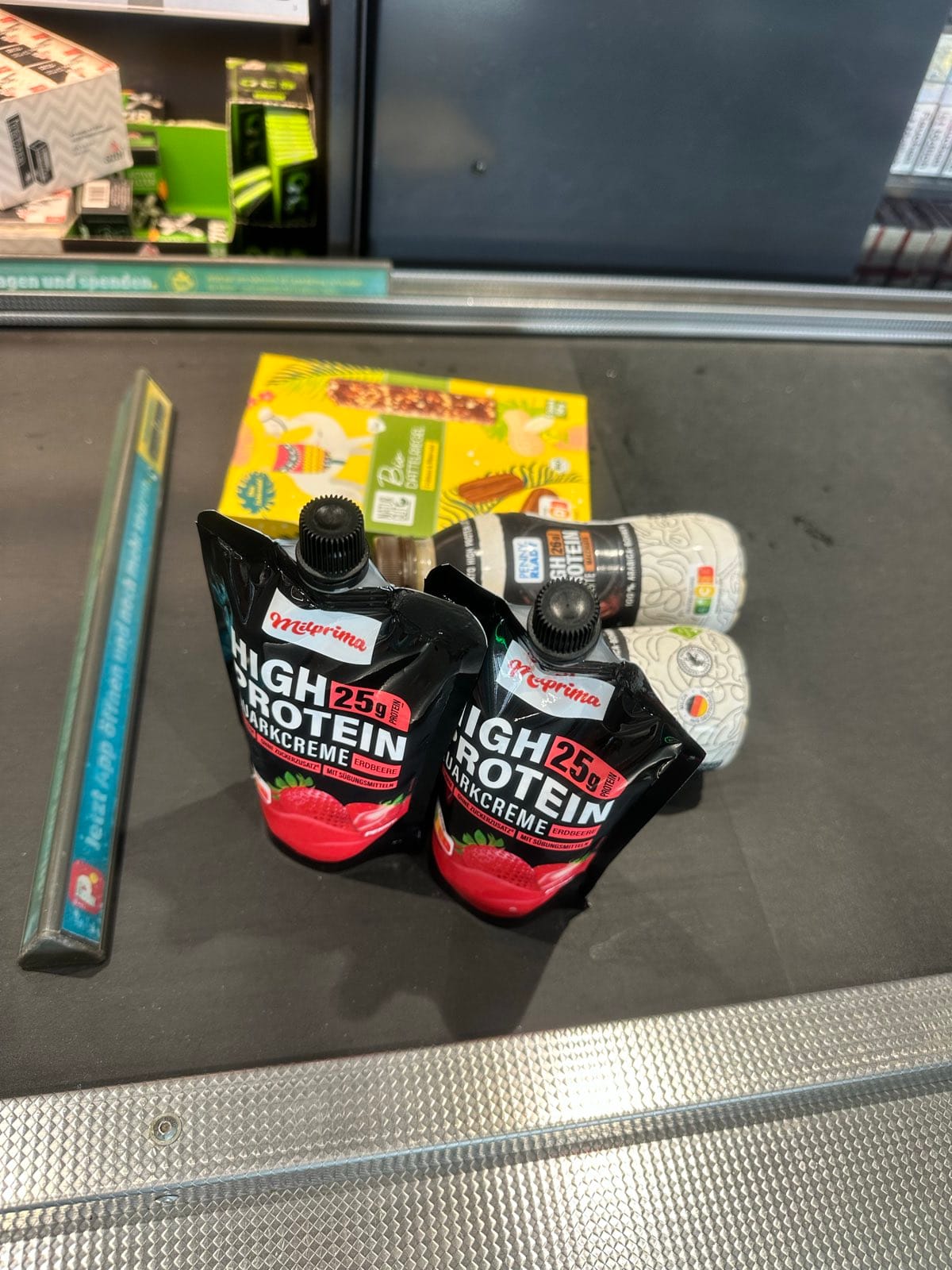 Two strawberry high protein pouches, a chocolate bar and two drinks on a German supermarket checkout belt