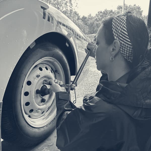 Evie beside TS2 with a spat removed, working a torque wrench on a wheel, water still beaded on the white paint