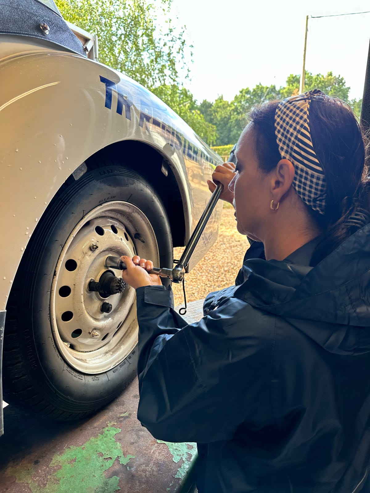 Evie beside TS2 with a spat removed, working a torque wrench on a wheel, water still beaded on the white paint