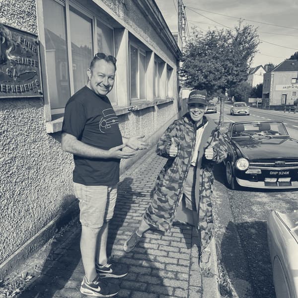 Evie on a French pavement in her camouflage dry robe and cap, both thumbs up, a black TR6 parked at the kerb behind