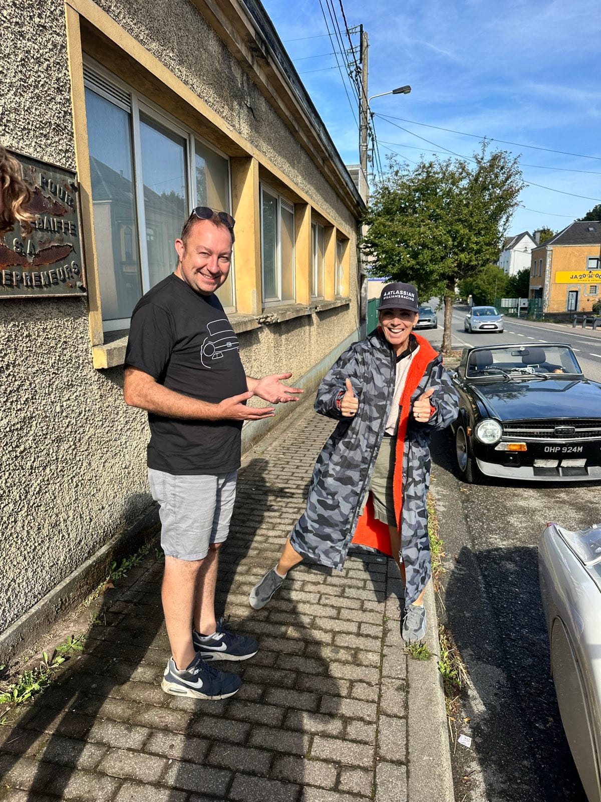 Evie on a French pavement in her camouflage dry robe and cap, both thumbs up, a black TR6 parked at the kerb behind