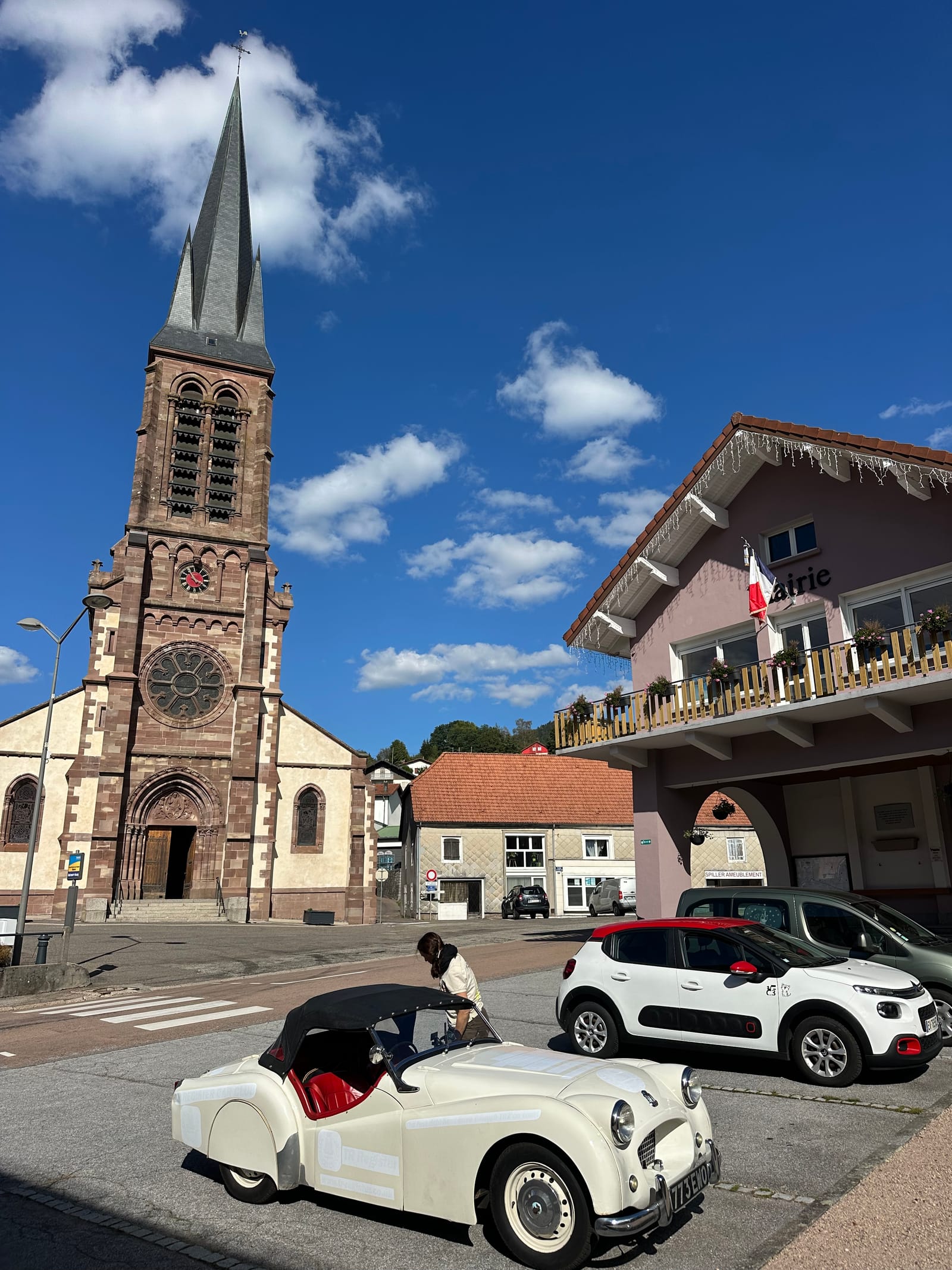 The red sandstone church at Saint-Maurice-sur-Moselle under a blue sky, TS2 parked at the kerb below and Evie climbing in