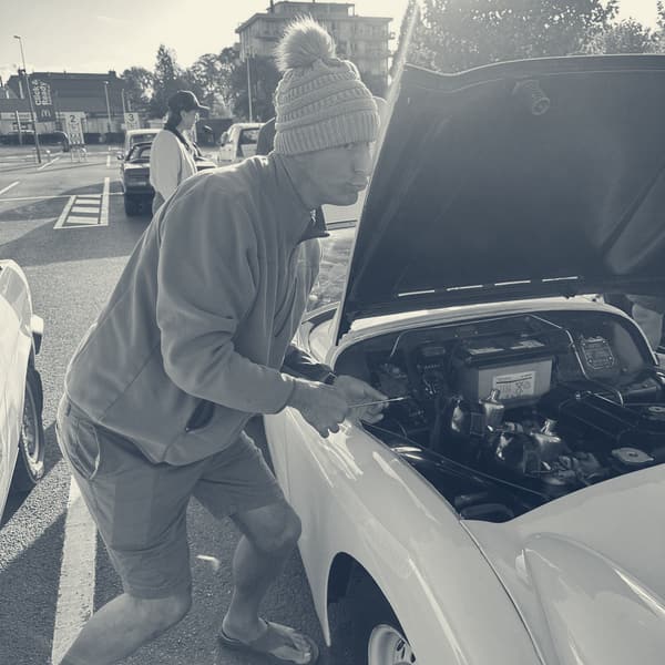A man in a bobble hat leaning over TS2's open bonnet in the car park, low sun flaring behind him