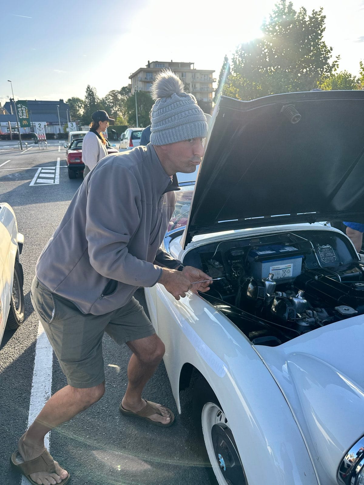 A man in a bobble hat leaning over TS2's open bonnet in the car park, low sun flaring behind him