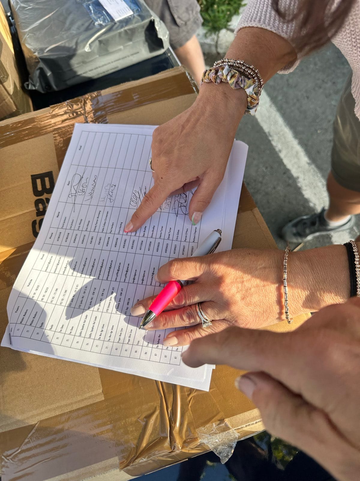 Two pairs of hands over the printed start list, one pointing at a line, the other holding a pen, the sheet resting on a cardboard box
