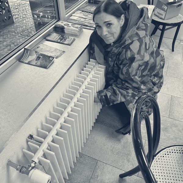 Evie crouched against a radiator under a service station window, both hands on it