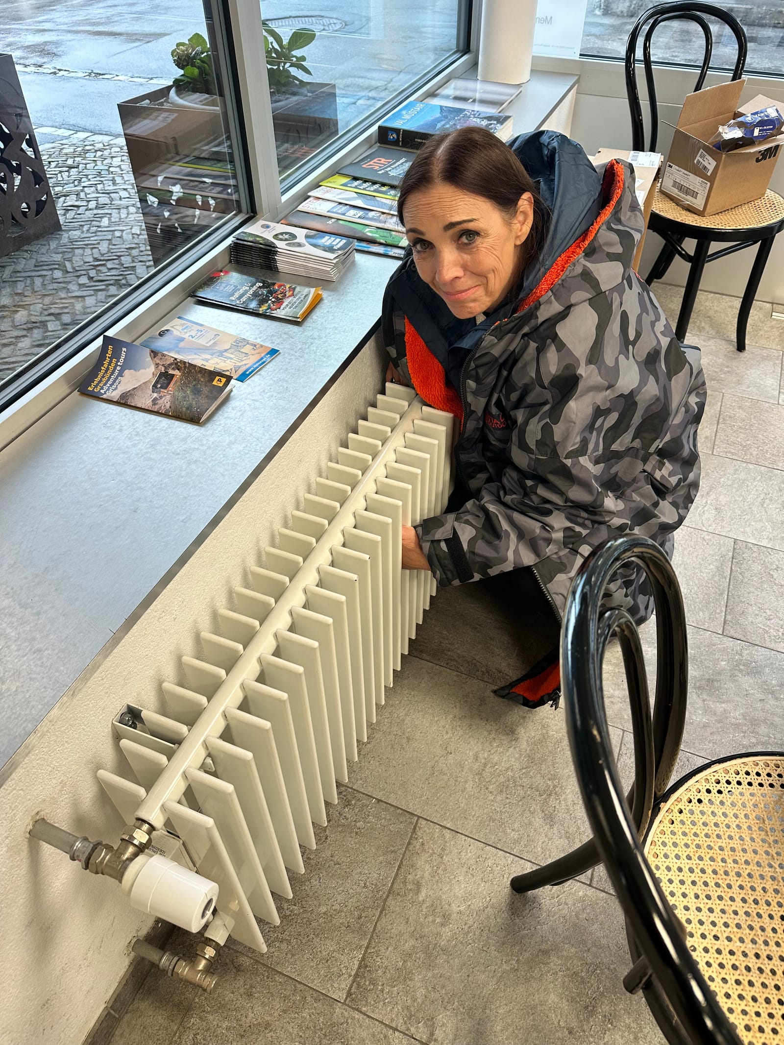 Evie crouched against a radiator under a service station window, both hands on it