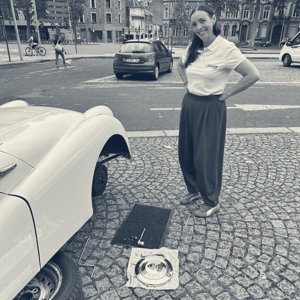 Evie standing hands on hips beside TS2 with her front wheel off, the hubcap and tools laid out on a cloth on the cobbles