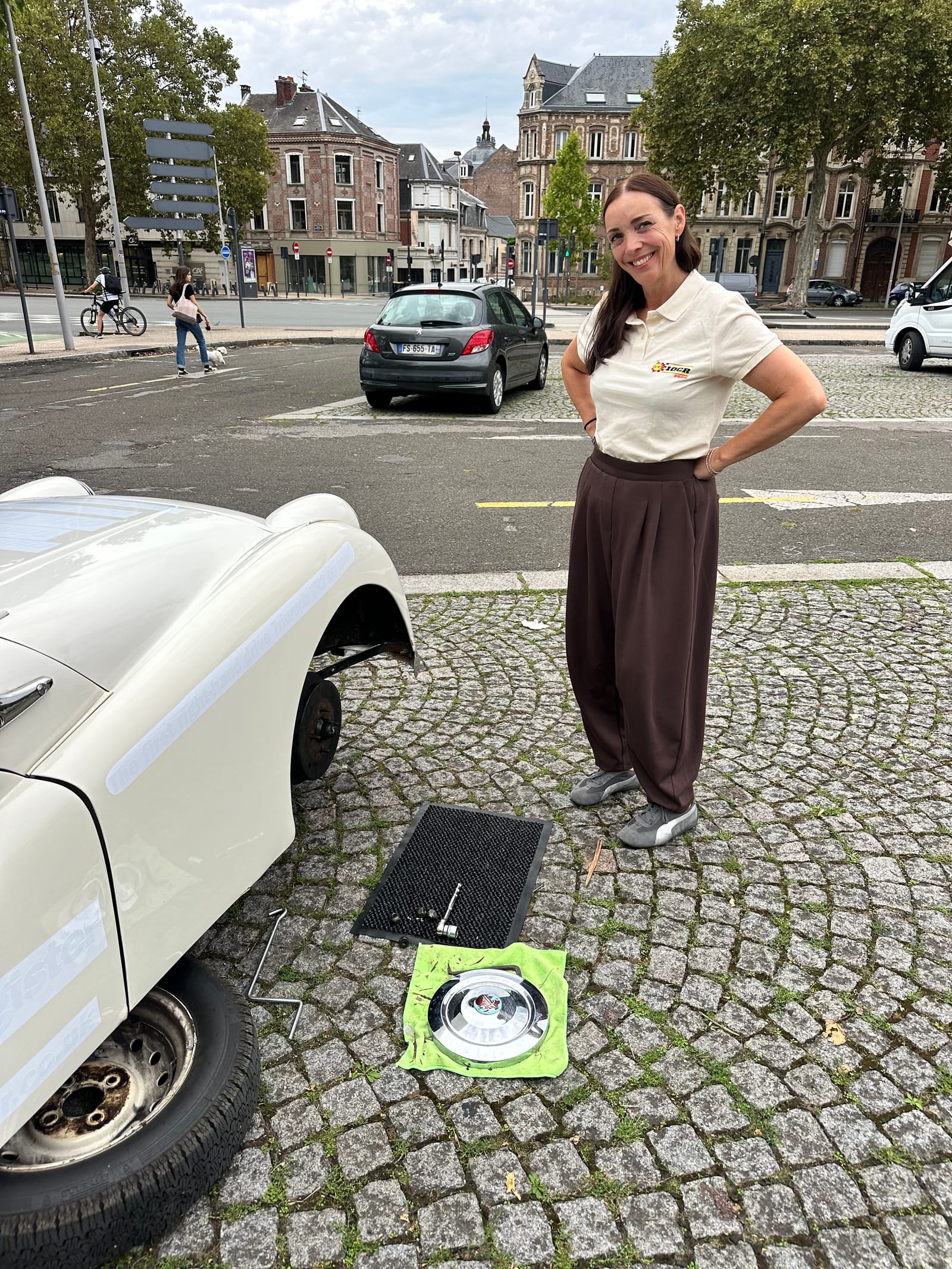 Evie standing hands on hips beside TS2 with her front wheel off, the hubcap and tools laid out on a cloth on the cobbles