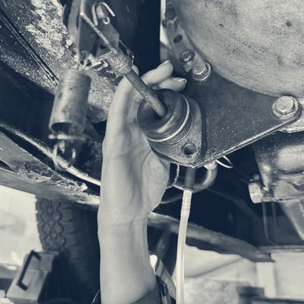 A hand reaching up to the clutch operating rod and its rubber boot on the underside of the gearbox bellhousing