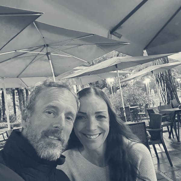 Evie and Andy at a café table under umbrellas in the evening, both smiling into the camera