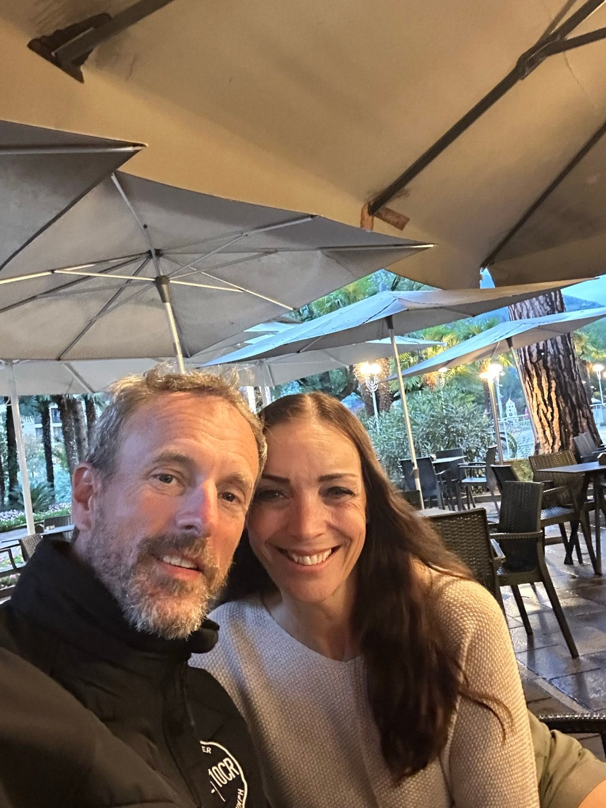 Evie and Andy at a café table under umbrellas in the evening, both smiling into the camera