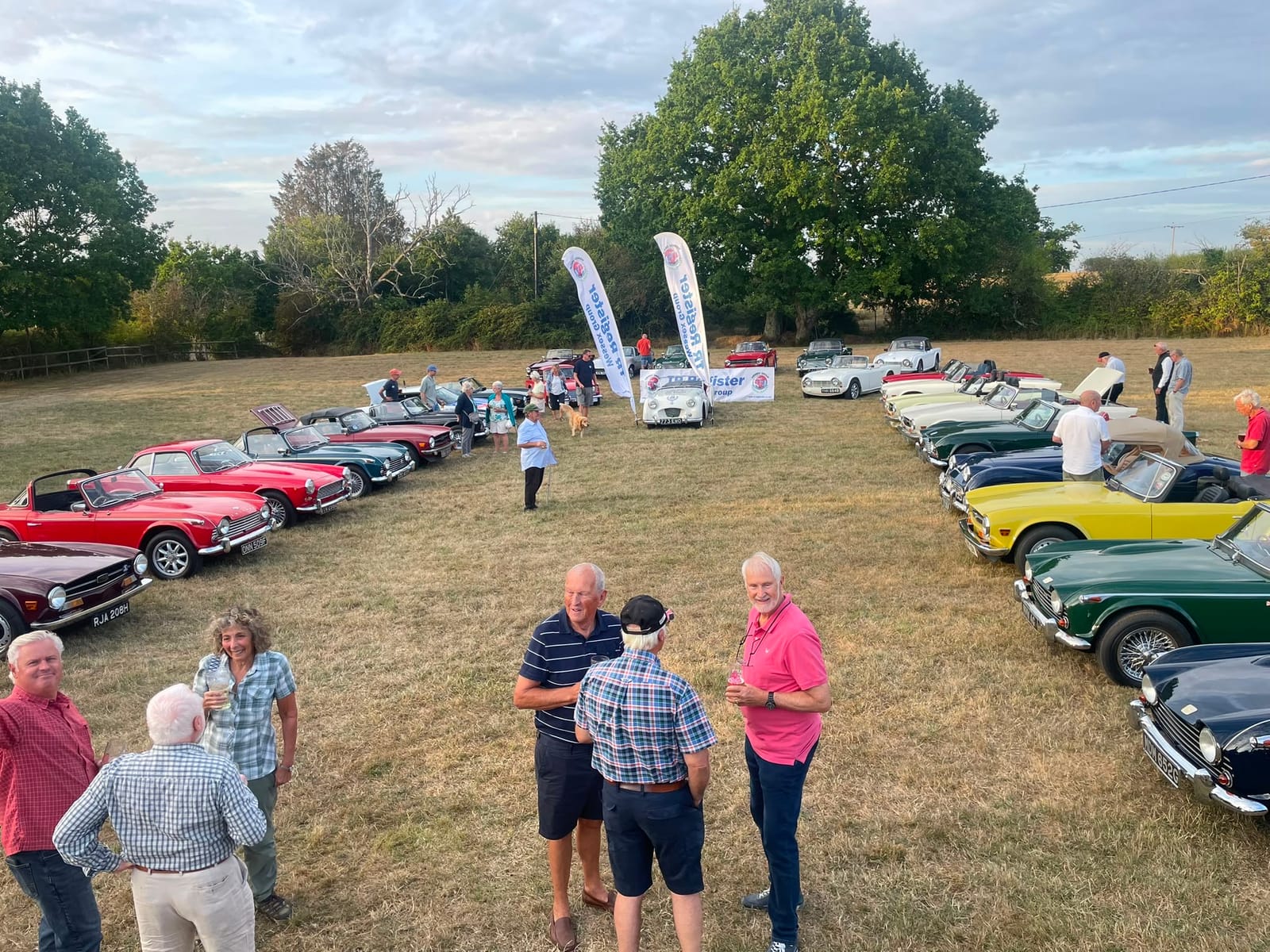 The same field later in the evening, members standing about in groups with drinks as the light goes
