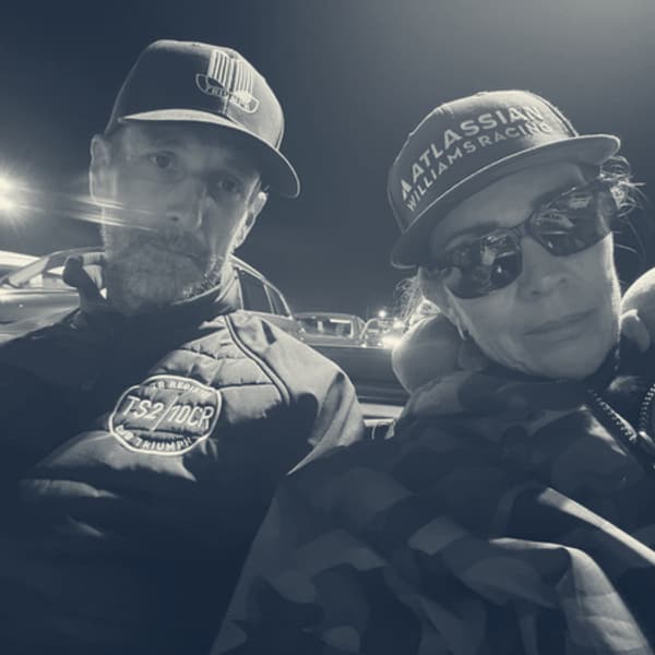 Andy and Evie in the ferry queue at night, both in caps, Evie in sunglasses, a red Triumph under the floodlights behind