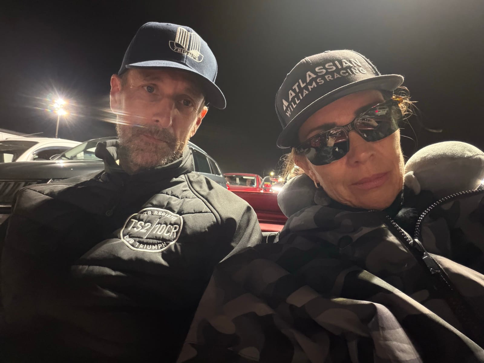 Andy and Evie in the ferry queue at night, both in caps, Evie in sunglasses, a red Triumph under the floodlights behind