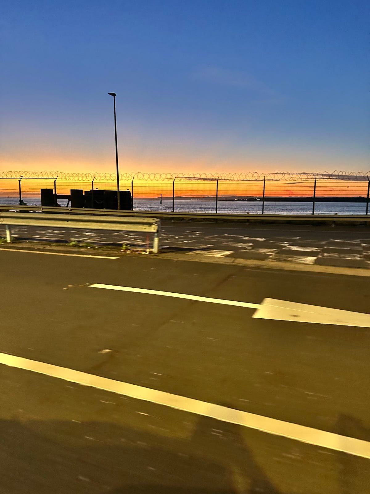 Sunrise over flat water behind a razor wire fence, seen from the road along the port