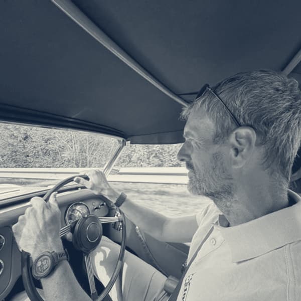 Andy at the wheel in a white polo shirt, hood up, sunlit trees through the windscreen