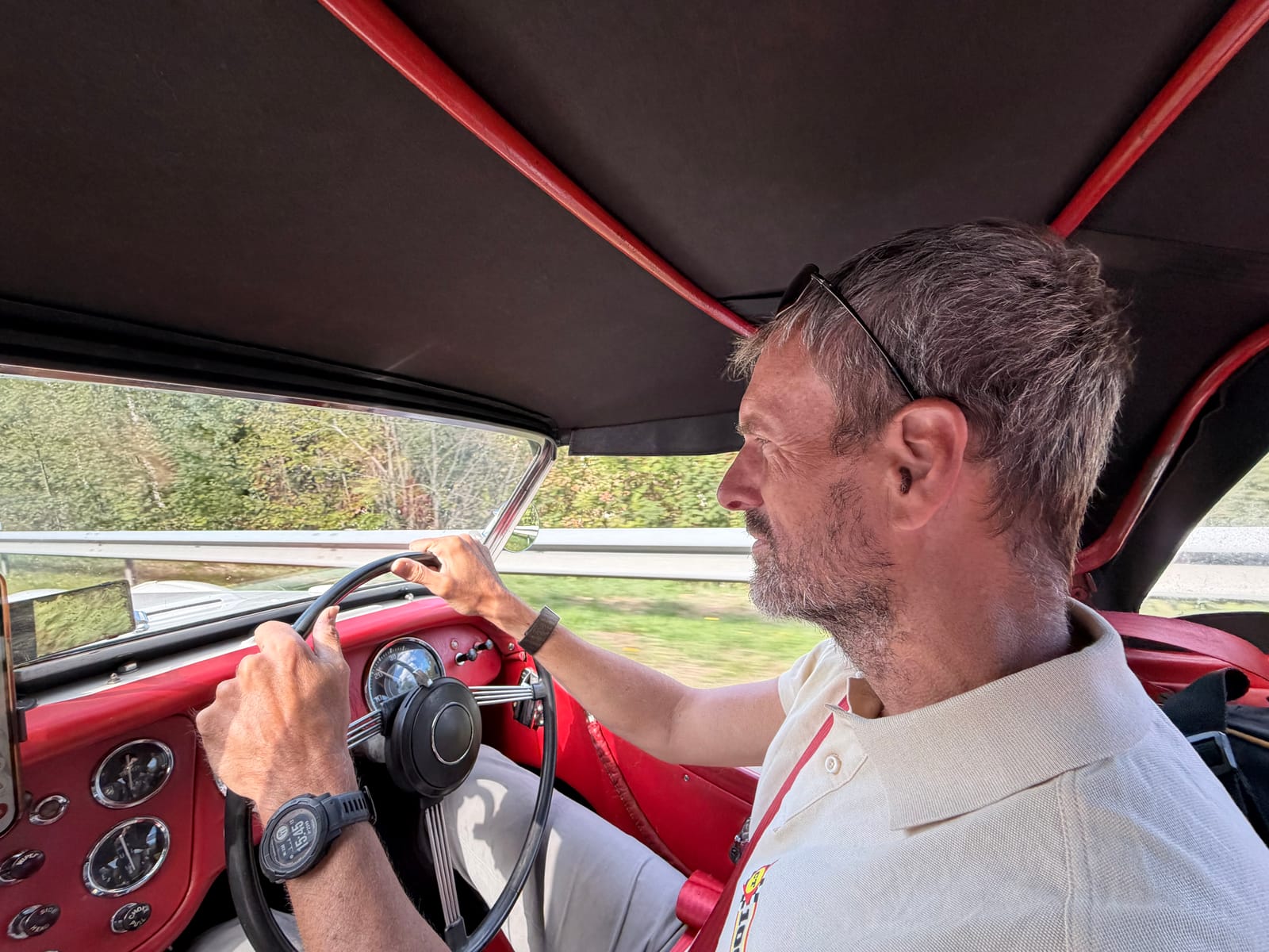 Andy at the wheel in a white polo shirt, hood up, sunlit trees through the windscreen