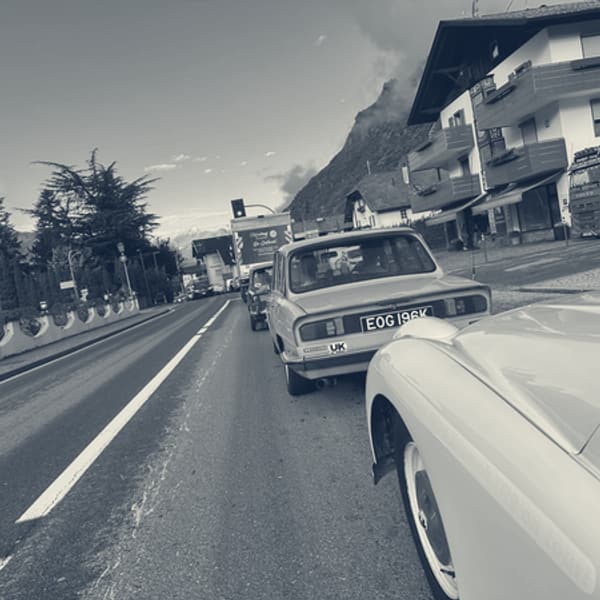 A pale blue Triumph 2000 saloon ahead of TS2 on the main road out of Merano, mountains behind the rooftops