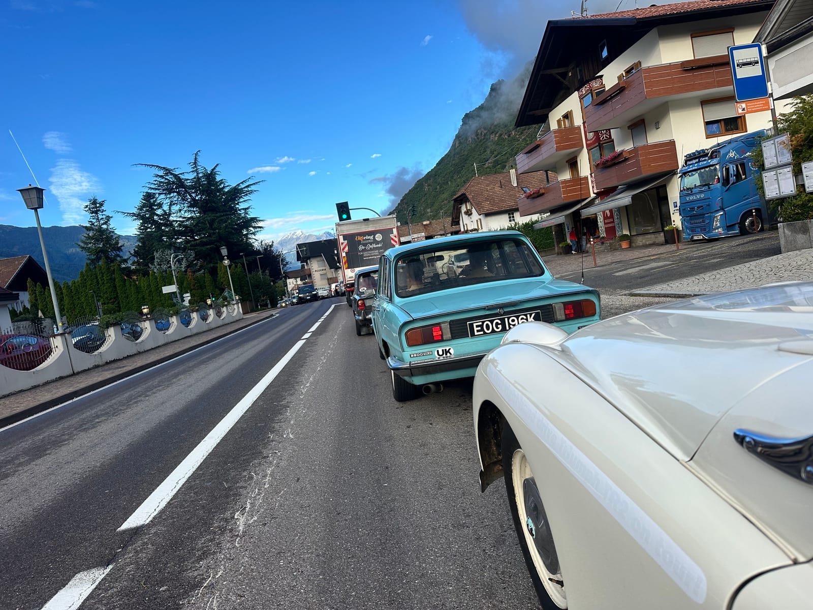 A pale blue Triumph 2000 saloon ahead of TS2 on the main road out of Merano, mountains behind the rooftops