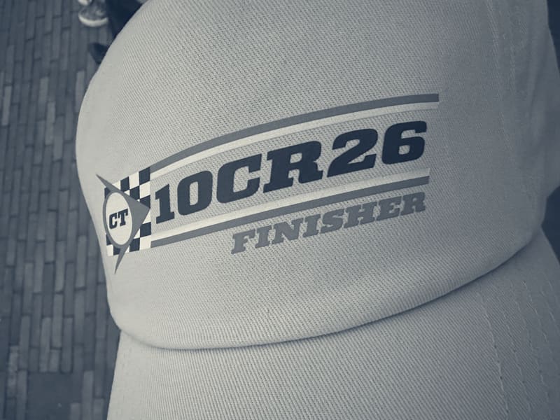 A grey Club Triumph cap with the 10CR26 Finisher badge, held over the cobbles.