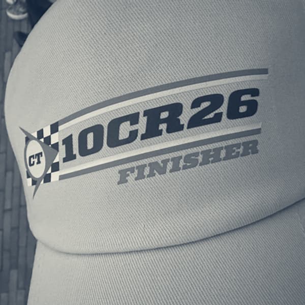A grey Club Triumph cap with the 10CR26 Finisher badge, held over the cobbles.