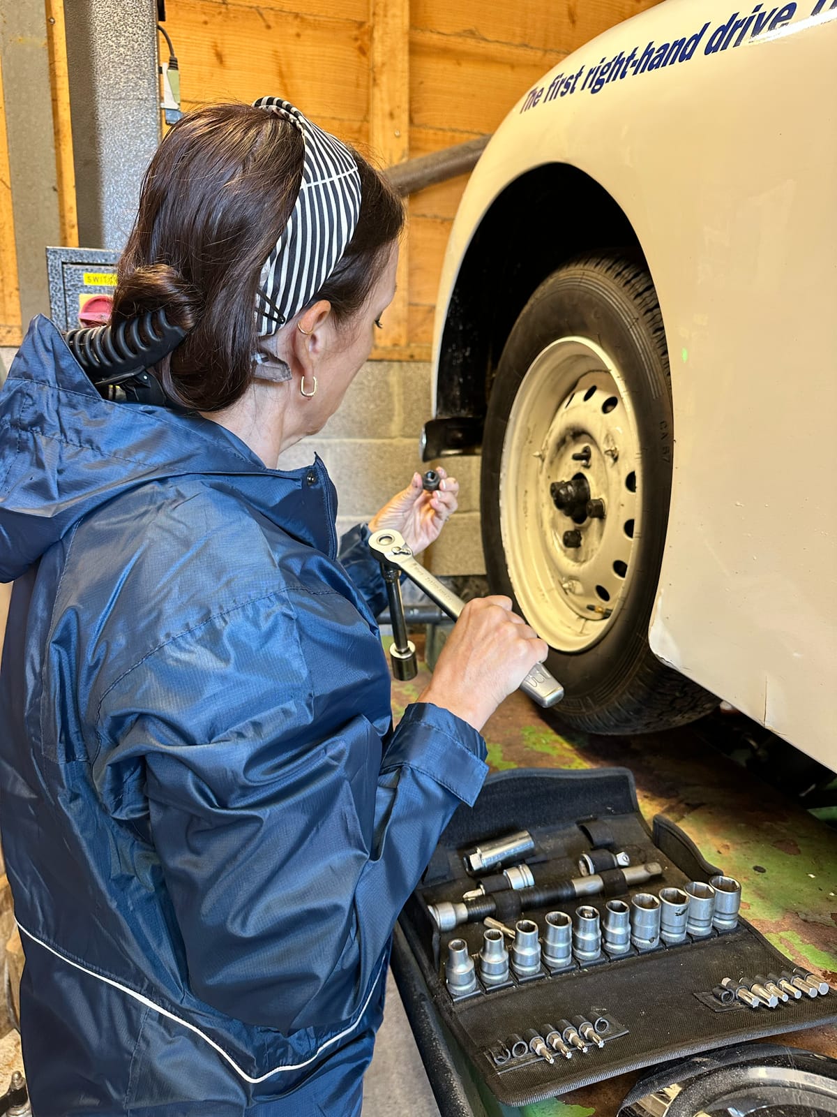 Evie at a wheel with a ratchet, a socket set laid open on the lift ramp beside her