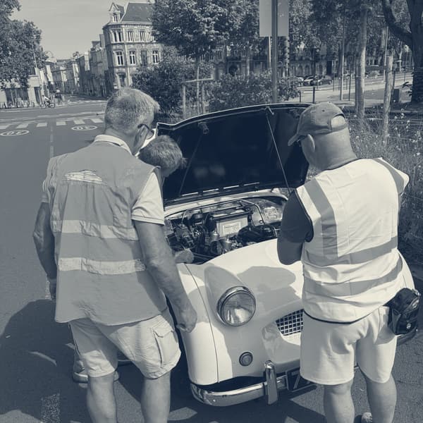 Three men in hi-vis vests peering into TS2's engine bay from the front, the bonnet up and a stone town house across the junction behind